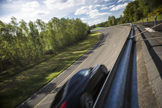 A dark car is captured from a high angle, possibly from a drone or elevated viewpoint, as it navigates a winding asphalt road through a lush, green landscape under a bright, partly cloudy sky. The road features clear markings and guardrails on the right.