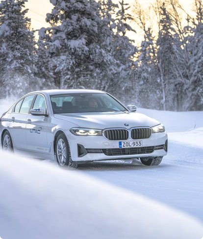 A white BMW sedan drives through a snowy landscape with snow-covered trees at sunset, kicking up snow from its tires.