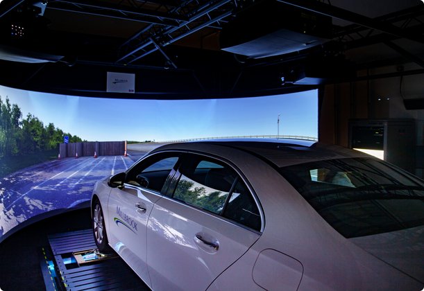 A white car is positioned inside a driving simulator with a large, curved screen displaying a realistic road landscape.