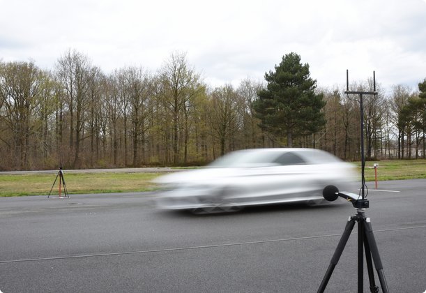 A blurry, fast-moving car on a test track, with a measurement microphone on a tripod in the foreground. Trees and a slightly cloudy sky are visible in the background.