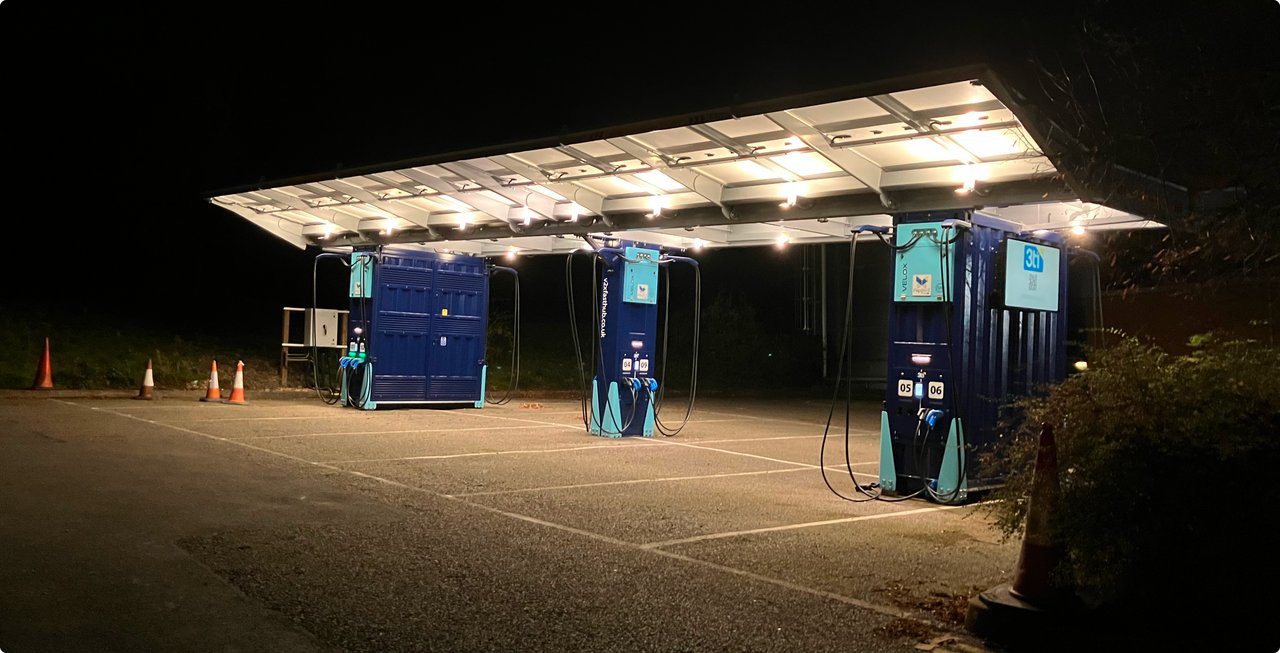 An electric vehicle charging station at night, featuring a canopy covered with brightly lit solar panels. Several blue charging pillars stand on a gravel lot. The surroundings are dark, highlighting the station's illumination.
