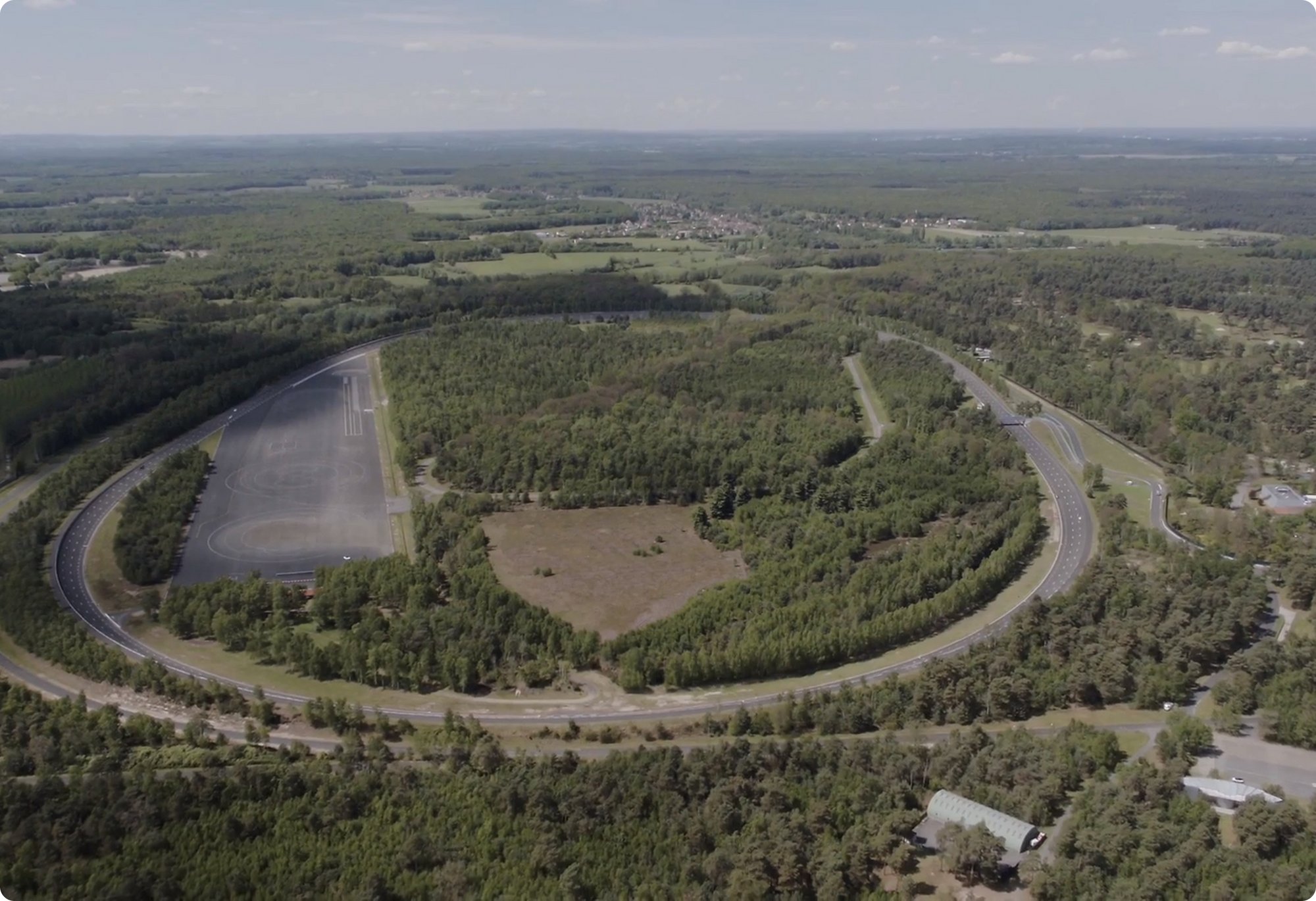 An aerial view of a large, U-shaped forested area, possibly a test track or a former industrial site, with a dark, open area at one end of the "U" and roads or clearings visible within and around the trees.
