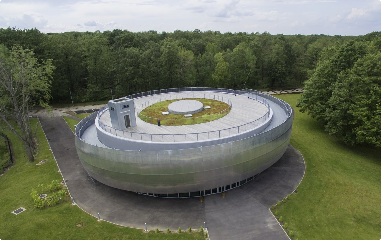 Aerial view of a modern, oval-shaped building with a green roof, surrounded by dense green trees.