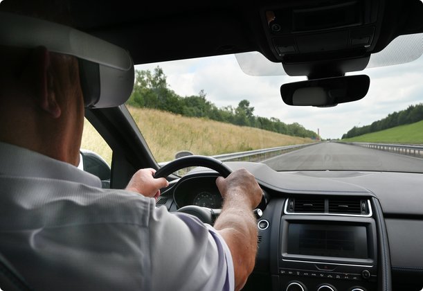 A driver, seen from behind, is at the wheel of a car on a test track that winds through a hilly landscape with grass and trees.