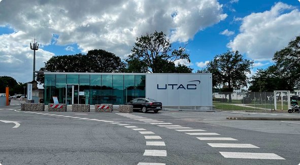 Le portail d'entrée moderne d'un site d'entreprise avec un poste de garde vitré et une enseigne affichant "UTAC" sous un ciel nuageux.