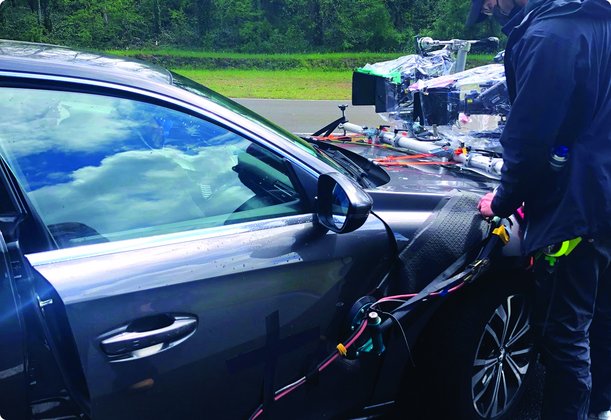 Eine Person steht neben einem dunklen Auto, das mit komplexer Kameraausrüstung auf dem Dach und an der Seite montiert ist, wahrscheinlich für Filmaufnahmen oder Datenerfassung. Die Spiegelung im Auto zeigt einen bewölkten Himmel.
