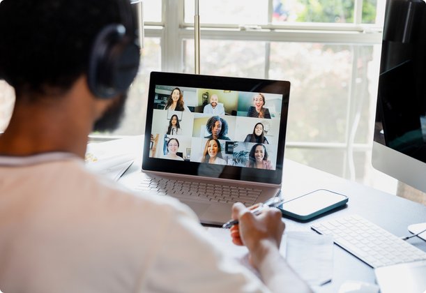 A person wearing headphones sits in front of a laptop, participating in a video conference call with multiple attendees. The person's back is visible in the foreground, while the laptop screen displays various faces.
