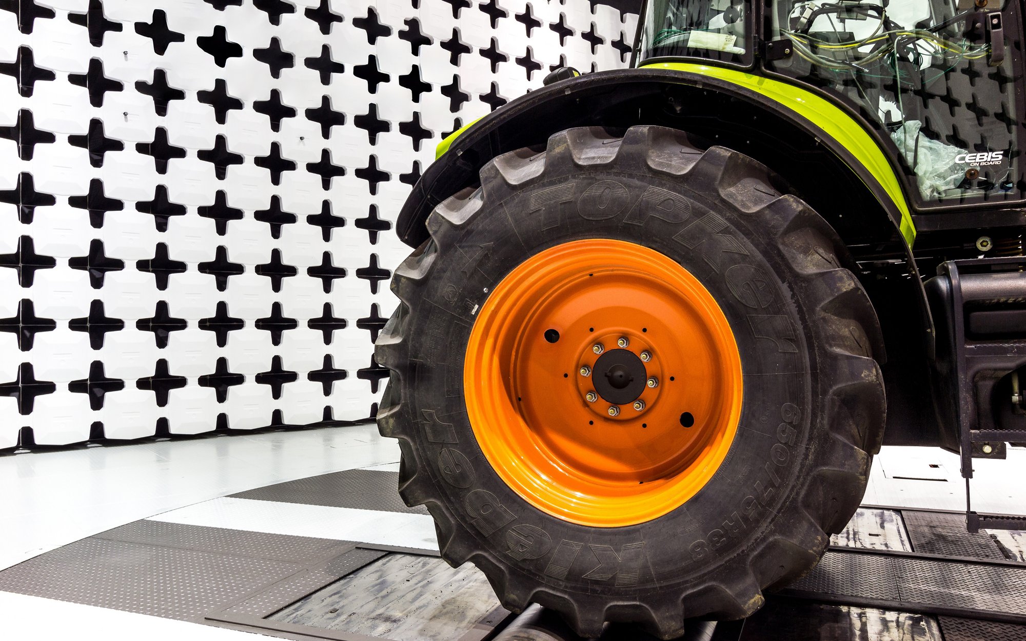 Close-up of a large tire with an orange rim from a tractor or similar agricultural machine. In the background, there is a wall with a black and white cross pattern, typically used for optical measurements.