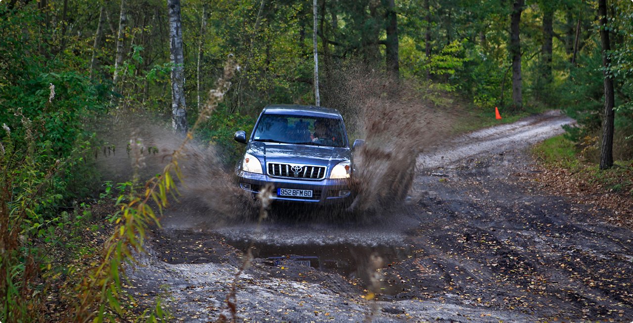 A dark SUV drives through a large muddy puddle on an unpaved forest road, creating a lot of splash.