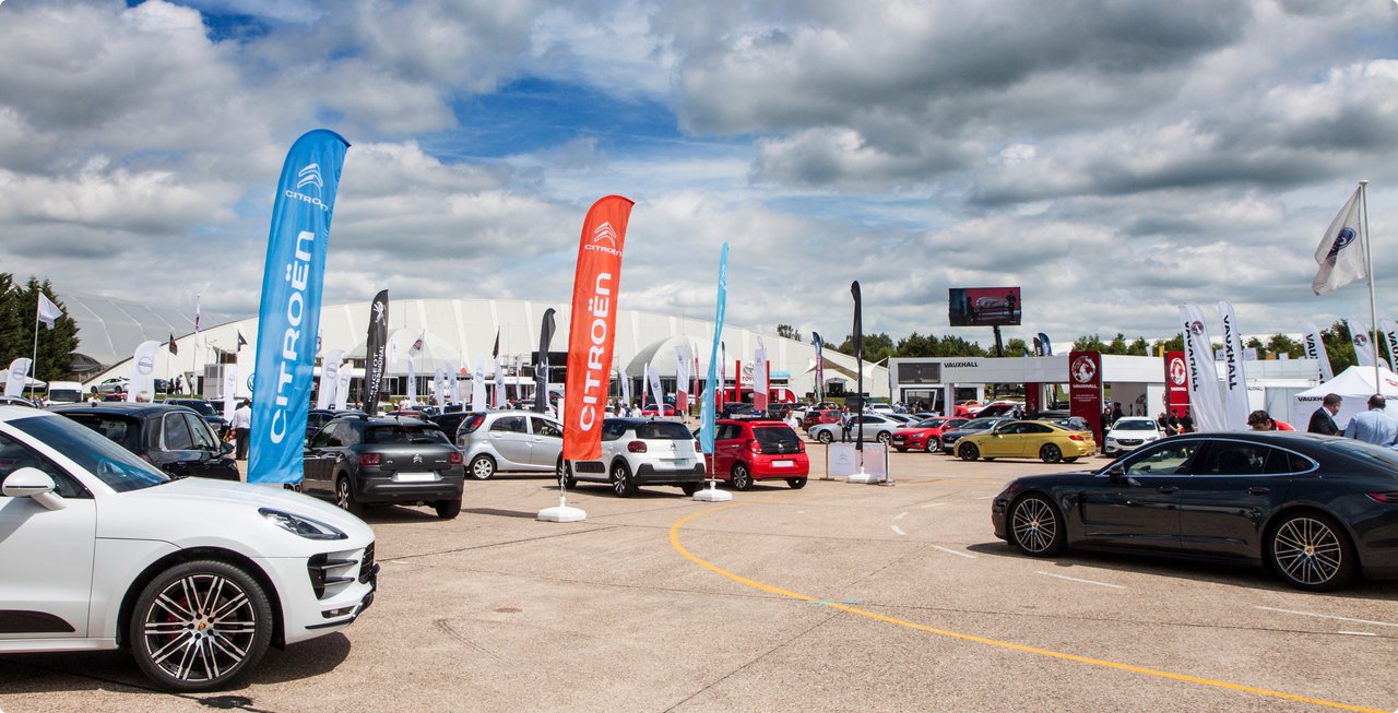 An outdoor view of an automotive event or fair, featuring several car models, tall promotional flags, and white tents on a gravel lot under a bright sky.