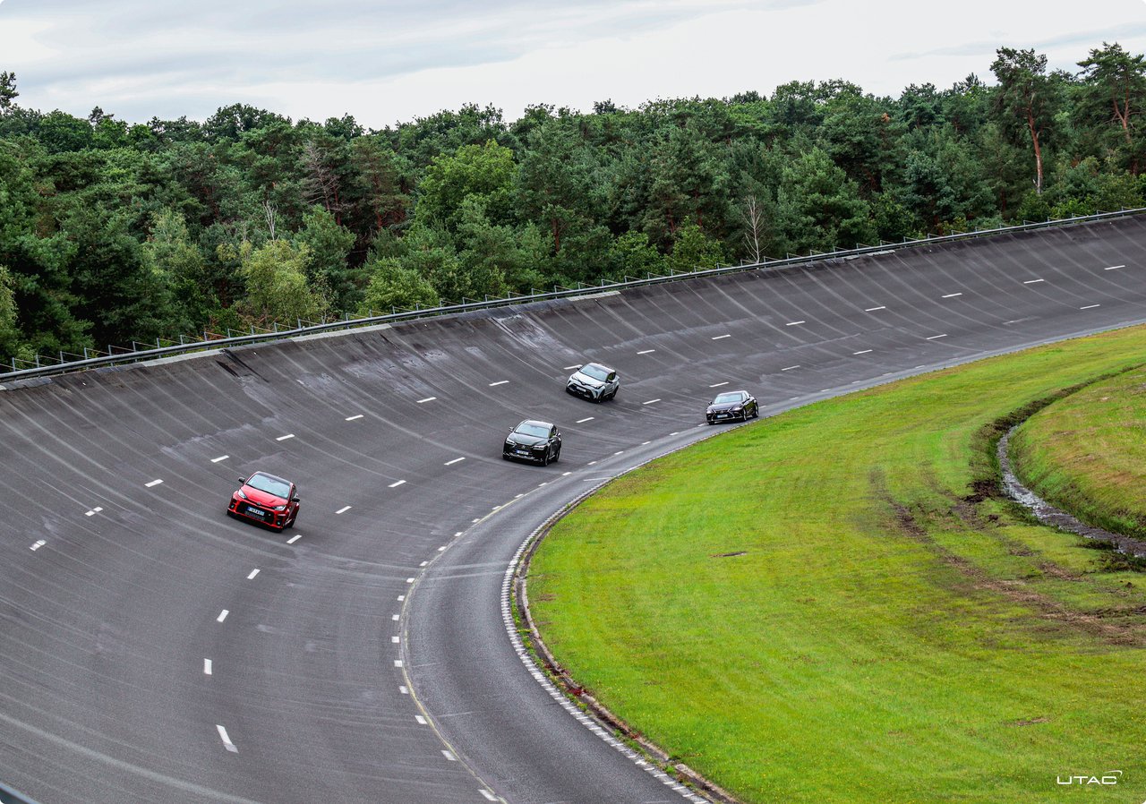 Several race cars, including a red vehicle in the foreground, are driving on an asphalt racetrack with a green curve and trees in the background.