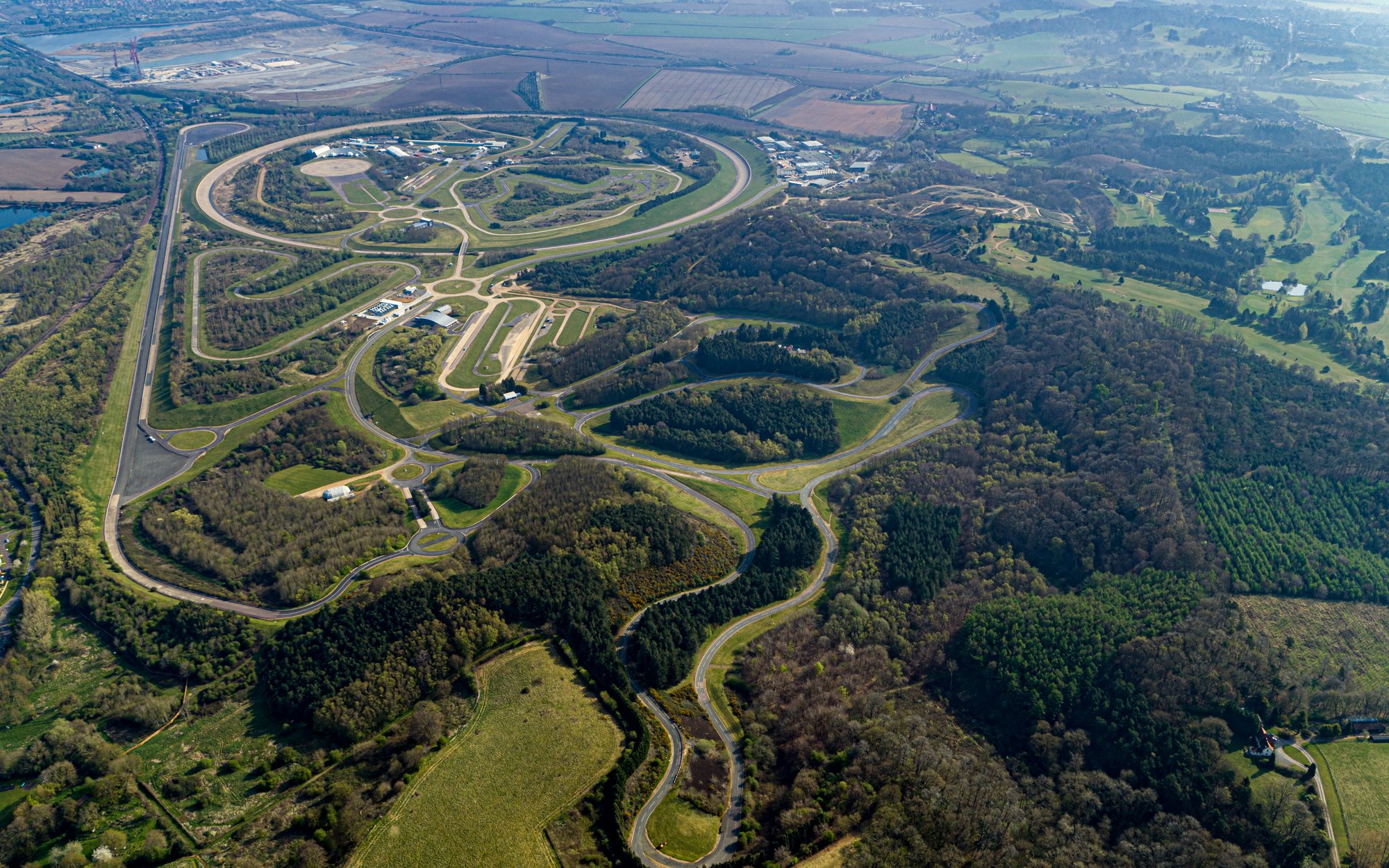 A sprawling aerial view of a test track facility with multiple oval and winding driving courses, surrounded by dense forests and green areas, under a clear sky.