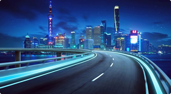Une autoroute urbaine illuminée de nuit, serpentant le long d'une rivière ou d'une baie, avec une ligne d'horizon brillamment éclairée en arrière-plan présentant des gratte-ciel et une tour proéminente.