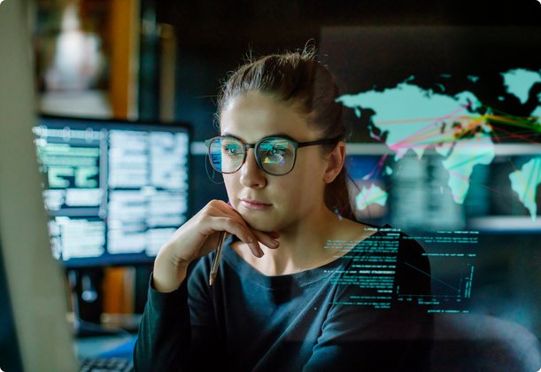 A focused woman with glasses sits in front of multiple screens, one of which displays a world map with data visualizations, suggesting data analysis or network monitoring.