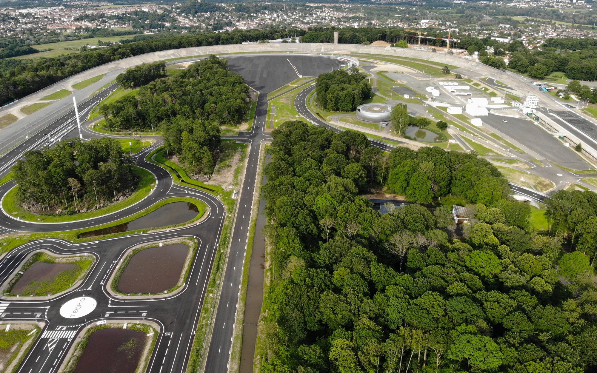 An aerial view of an extensive driving safety or testing facility with various tracks, roundabouts, and practice areas, surrounded by dense green trees and vegetation. In the background, settlements and further green areas can be seen.
