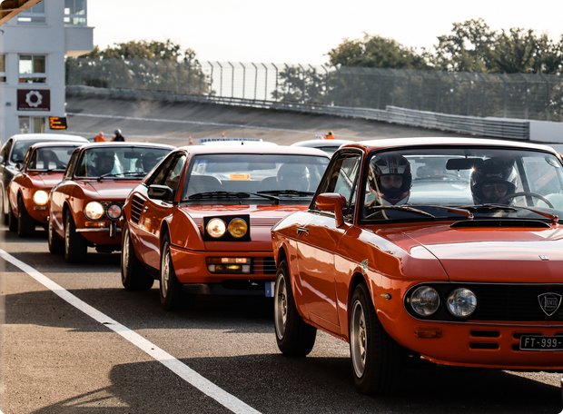 A line of several identical or very similar red vintage sports cars, possibly Lancia Fulvias, lined up on a race track or at an event. The cars have their headlights on.