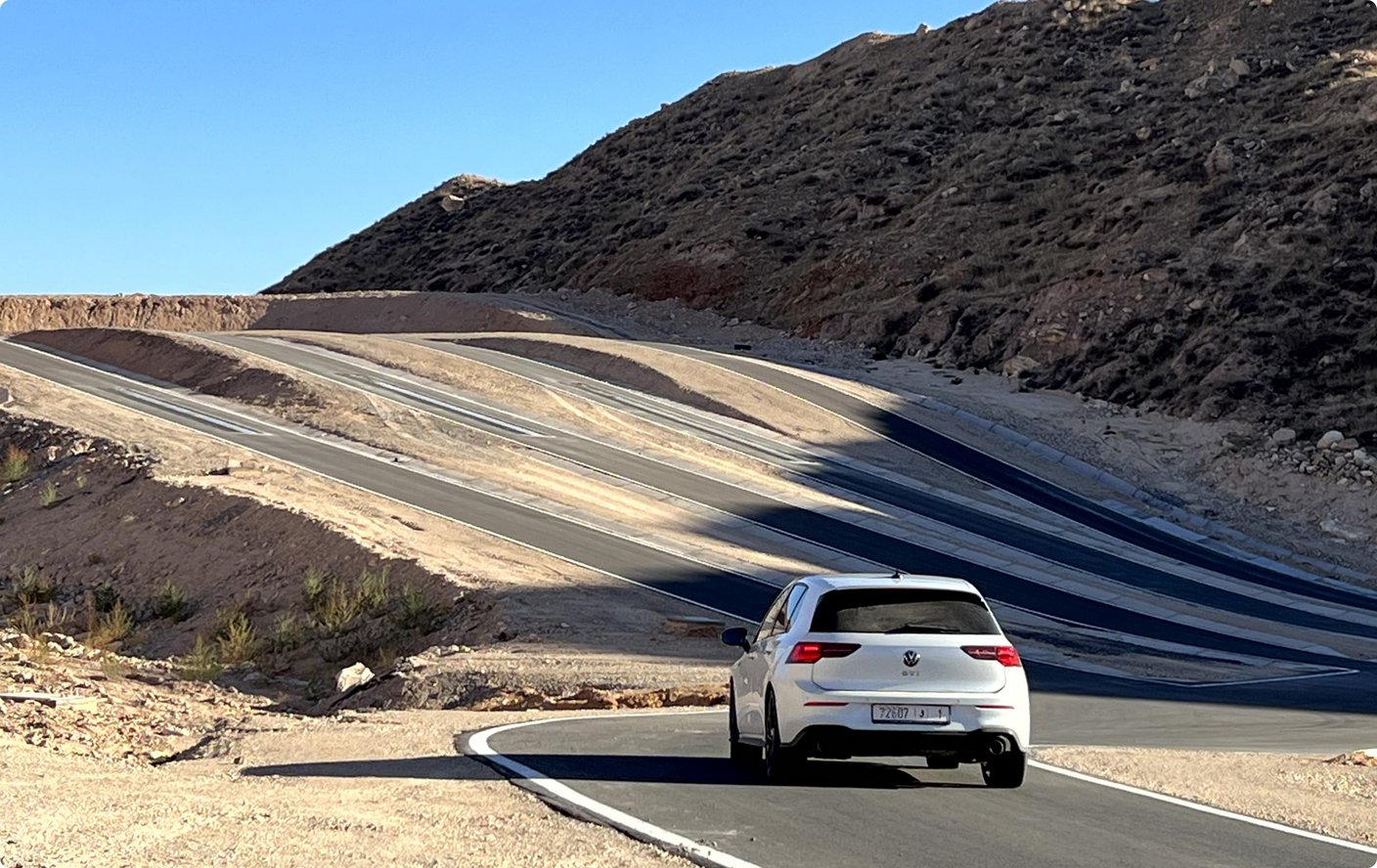 A white car drives on an asphalt road winding through a barren, mountainous landscape. The slopes show visible layers of rock and erosion.