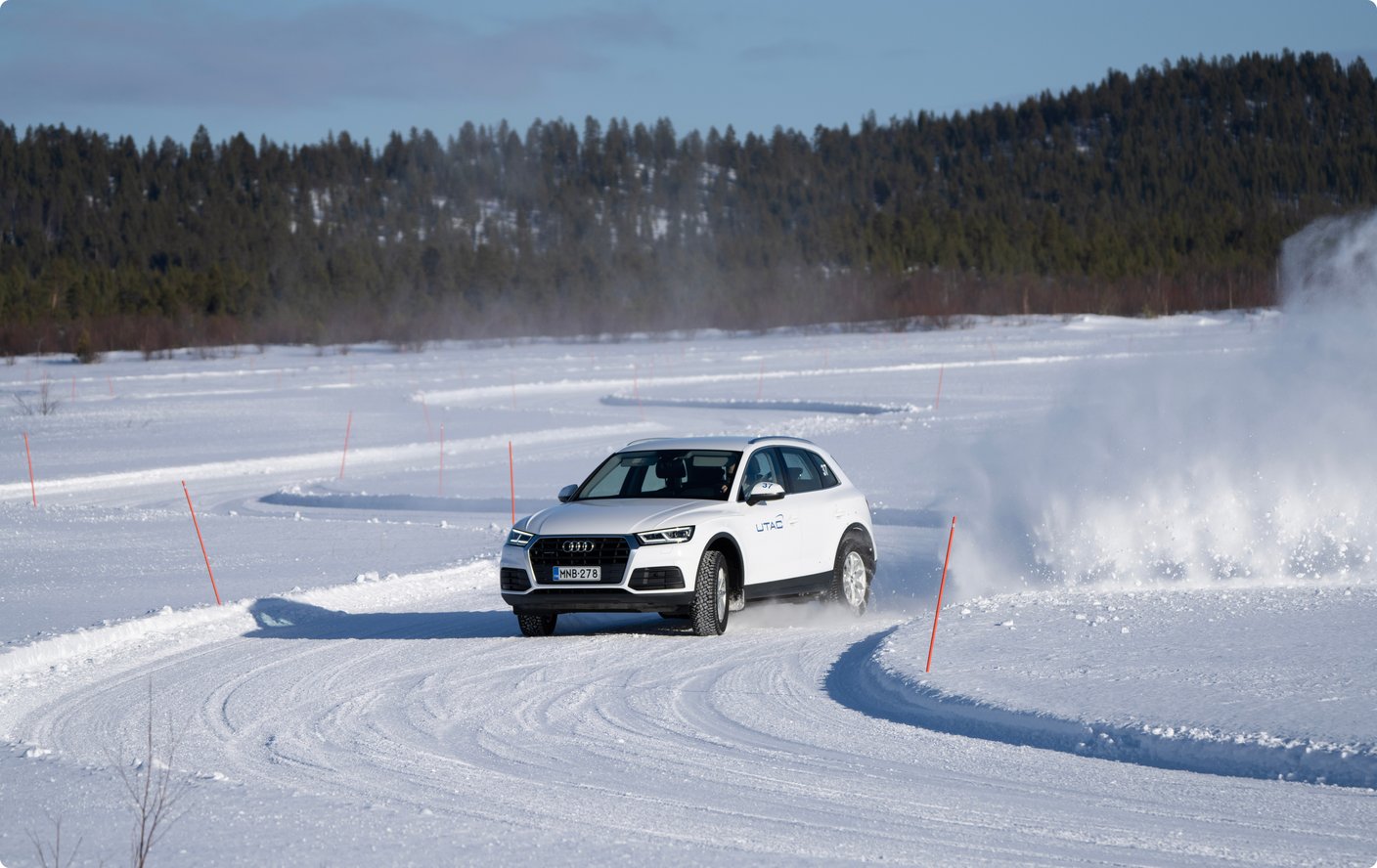 A white SUV drives on a snow-covered test track or frozen lake, kicking up snow. In the background, snowy forests are visible under a clear sky.