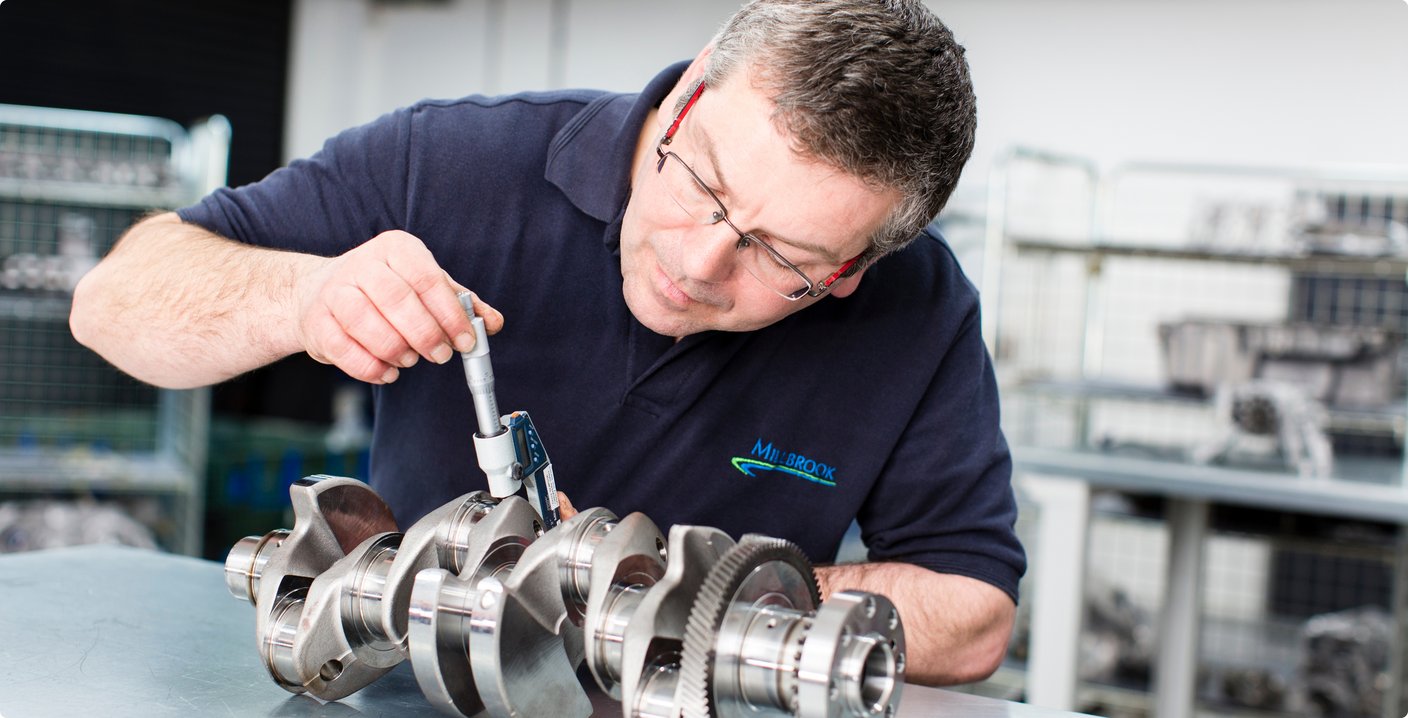 A bespectacled mechanic or technician in a dark polo shirt precisely measures a crankshaft with a caliper or micrometer. He is focused on measuring the component, which rests on a table in front of him.