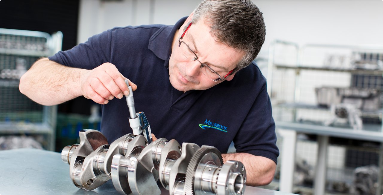 A bespectacled mechanic or technician in a dark polo shirt precisely measures a crankshaft with a caliper or micrometer. He is focused on measuring the component, which rests on a table in front of him.