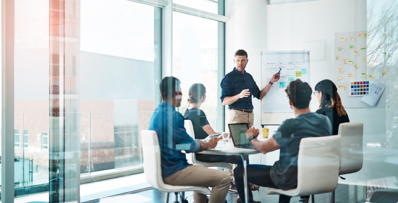 Employees in a modern office during a presentation; one person stands in front of a whiteboard explaining a concept.
