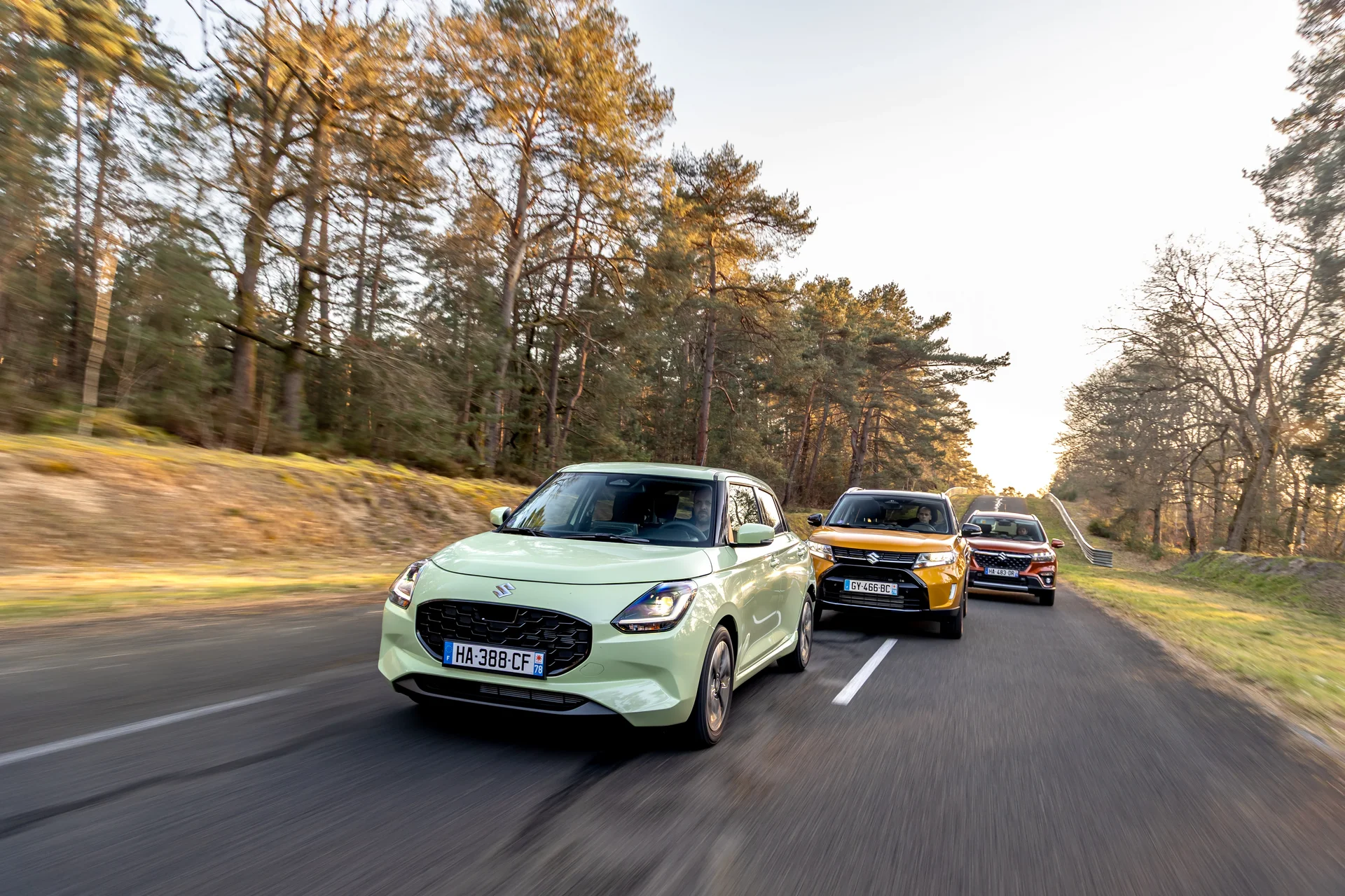 Three modern, compact electric vehicles driving in a line on an asphalt road through a forest or a tree-lined avenue. The leading vehicle is light green, followed by a yellow one and a darker vehicle. The scene is surrounded by trees and illuminated by warm sunlight, suggesting a test drive or convoy.