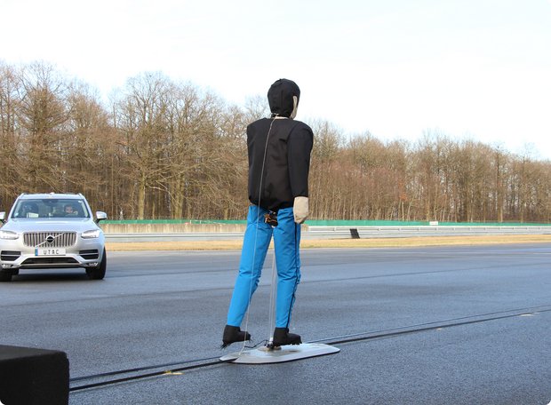 A test scene on a road, showing a silver SUV rolling towards a stationary test dummy, seen from behind, wearing a black torso and blue trousers. The scene takes place outdoors with trees in the background.