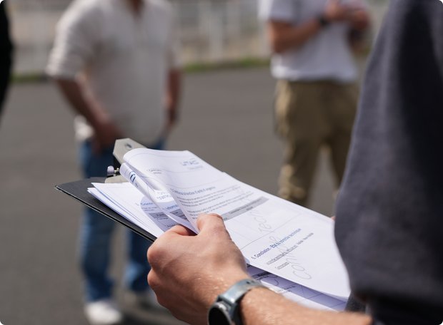 Close-up of a person holding a clipboard with notes, with other individuals standing in the blurred background.