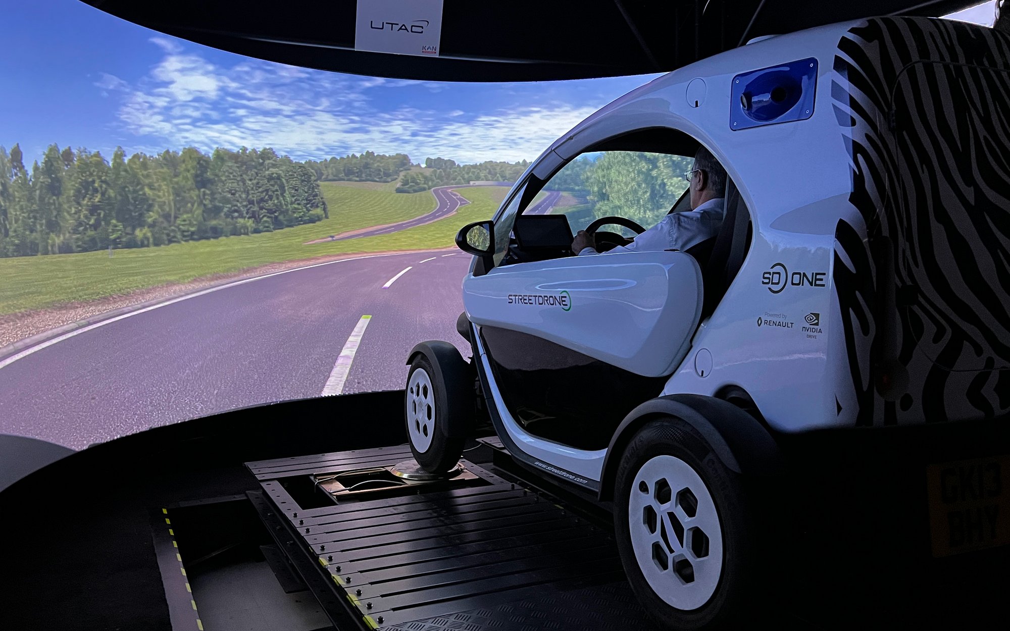 A photograph of a driving simulator, featuring a small, urban electric vehicle on a platform in front of a large, curved screen. The screen displays a rural road with trees, simulating a realistic driving environment. The vehicle has text like "StreetDrone" and logos from Renault and NVIDIA.