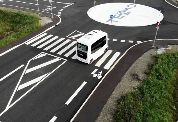 An autonomous white mini-bus drives on a crosswalk through a roundabout with a painted logo in the center.