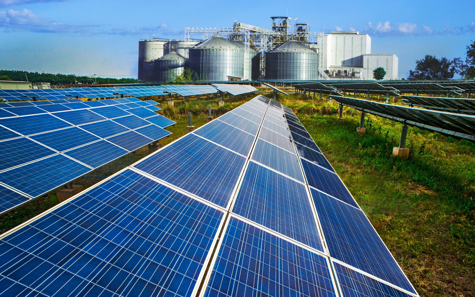 Solar panels in the foreground of a field, with an industrial facility featuring silos visible in the background under a blue sky.