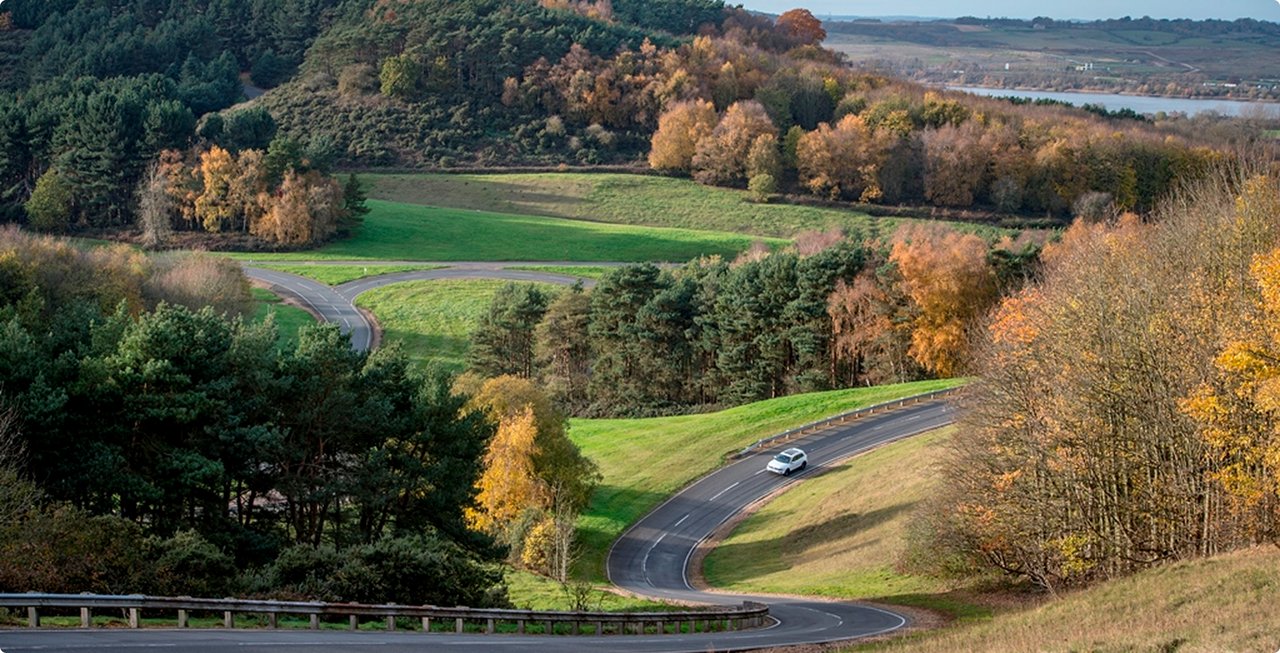 Autumnal landscape with winding roads, colorful trees, and a white car. A body of water or valley is visible in the background.