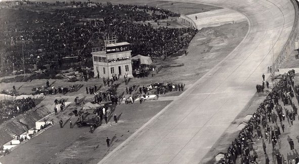 Une vue aérienne historique en noir et blanc d'un circuit de course avec des tribunes, une tour et des foules de personnes rassemblées, attendant un événement.