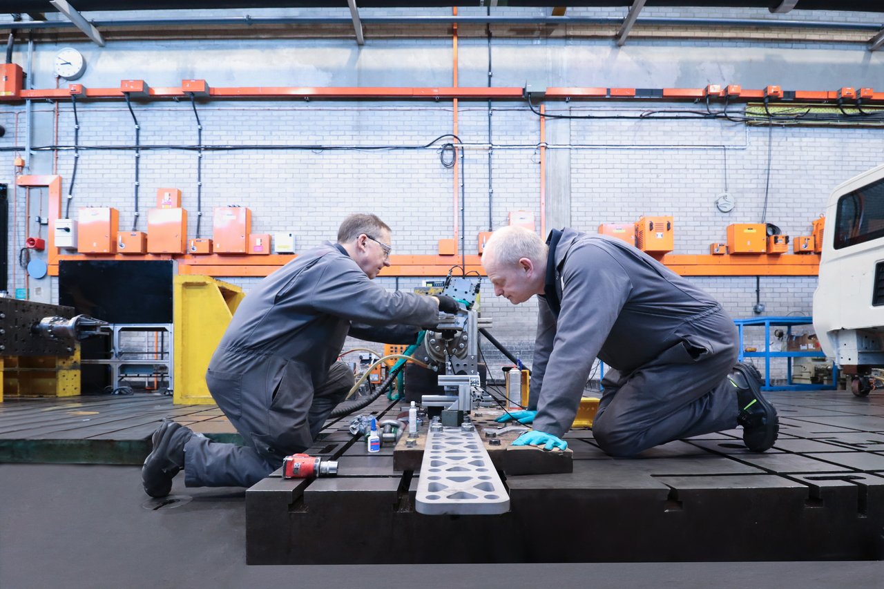 Two technicians in a workshop are kneeling and working on a piece of machinery or equipment on a workbench.