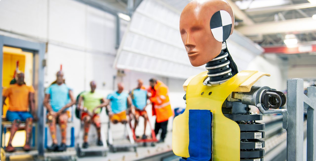 Close-up of a crash test dummy in the foreground, dressed in yellow and blue protective gear with a black and white target square on its head. In the blurred background, other dummies in various positions and colors are visible, some appearing to be on rolling carts.