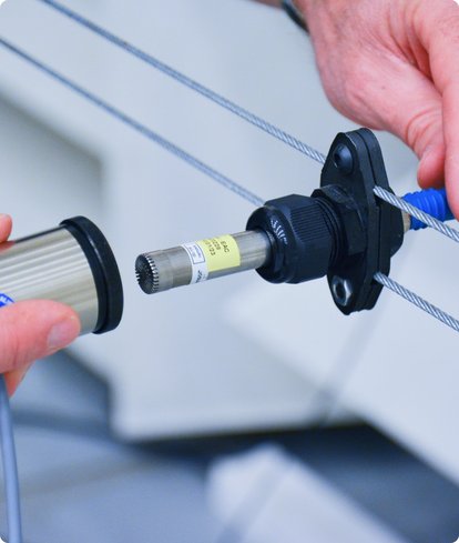 Close-up of hands connecting two parts of a measuring device or sensor. One part is silver-colored, the other is black with blue cables.