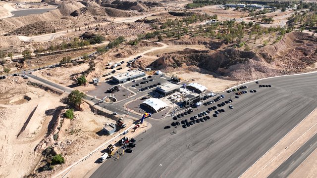 An aerial view displays an asphalt racing track with multiple curves, surrounded by barren, brown landscape and some trees. In the center of the facility, there is an area with a park-like design or possibly parked vehicles.