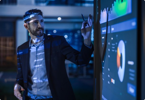 A bearded man in a suit stands in front of a large digital screen, pointing with a pen at complex diagrams and data, surrounded by a futuristic, blue-lit ambiance.