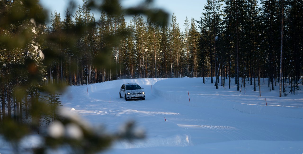 A white vehicle, possibly an SUV or pick-up truck, drives on a snow-covered road winding through a dense forest of evergreen trees. The foreground has blurred branches, suggesting a natural framing effect.