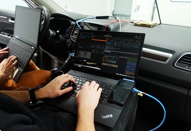 Close-up of two people working on laptops inside a car, seemingly connected to the vehicle to perform tests or data analysis.
