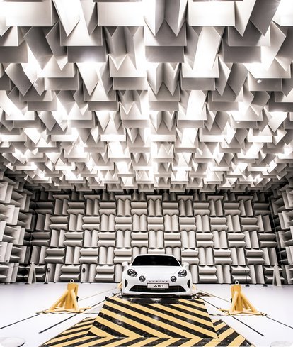A frontal view of a white car parked on a yellow and black striped surface in an anechoic chamber. The walls and ceiling of the room are covered with grey, sound-absorbing wedges.