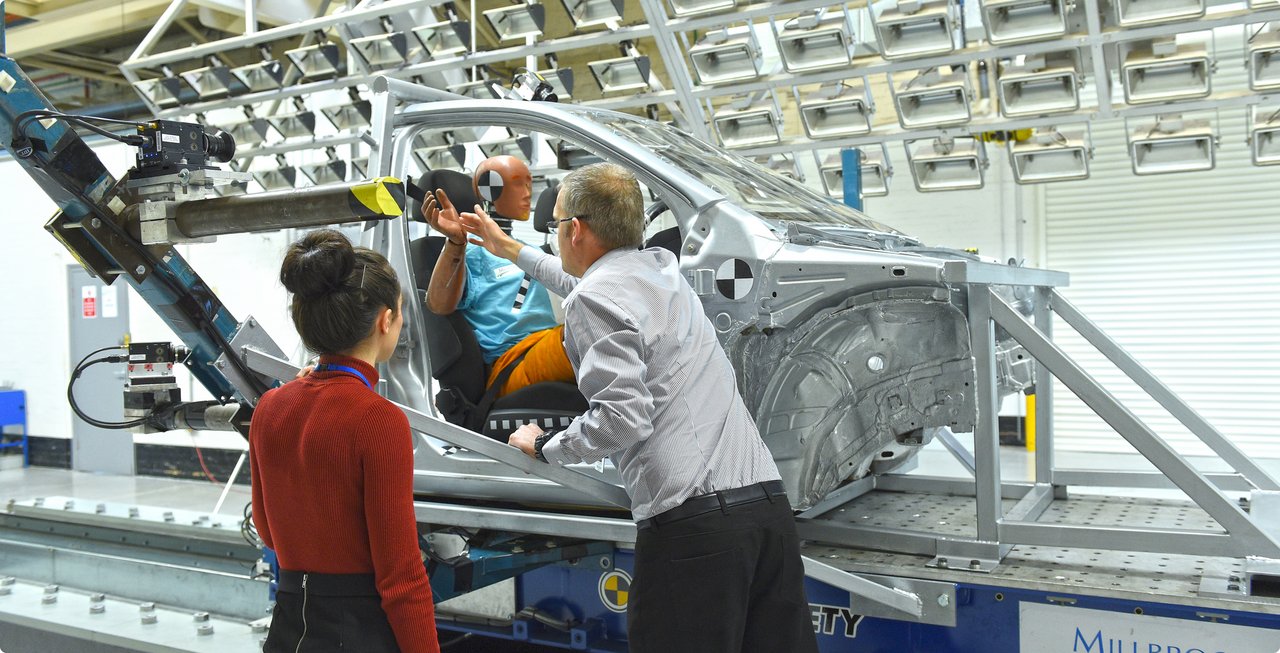 Two technicians, a woman in the foreground and a man sitting in a partially assembled car, inspecting components of a car body mounted on a test rig. Racks with testing equipment are visible in the background.