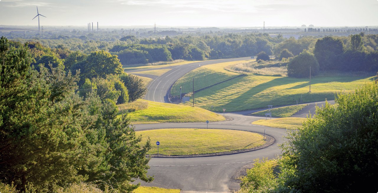Aerial view of a winding gravel track snaking through a landscape of trees and dry grasses. Distant buildings are visible on the horizon.