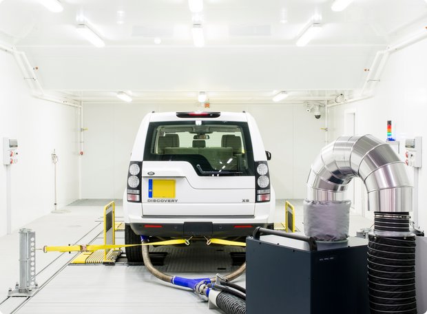 A white SUV is positioned on a dynamometer in a clean, white test chamber with exhaust extraction pipes, indicating emissions or performance testing.
