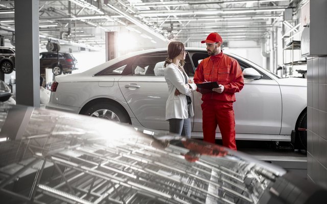 A mechanic in red work attire and a female client in a white coat are standing next to a silver car in an auto repair shop, discussing repairs.