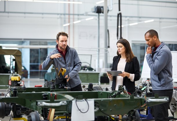 Engineering team in a production hall discussing technical details of a vehicle frame; one person holding a tablet.
