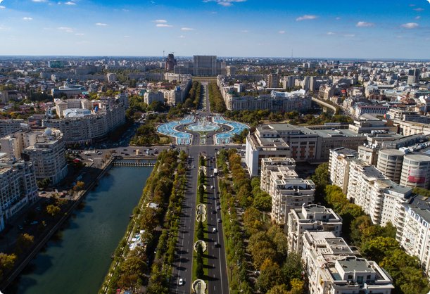 Vue aérienne d'un large boulevard bordé d'arbres traversant une ville dense, menant à un grand monument ou bâtiment circulaire orné au loin, avec une rivière ou un canal parallèle sur la gauche. Le paysage urbain présente de nombreux bâtiments résidentiels et commerciaux sous un ciel bleu clair.