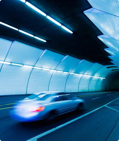 A blurred, silver car drives through a futuristically lit tunnel with bluish light. The tunnel lights are reflected on the car.