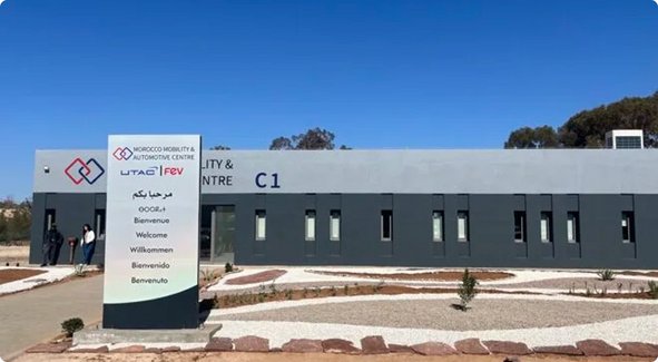 Un bâtiment moderne de plain-pied avec une façade grise et un grand panneau affichant plusieurs logos et noms d'entreprises, sous un ciel bleu clair.
