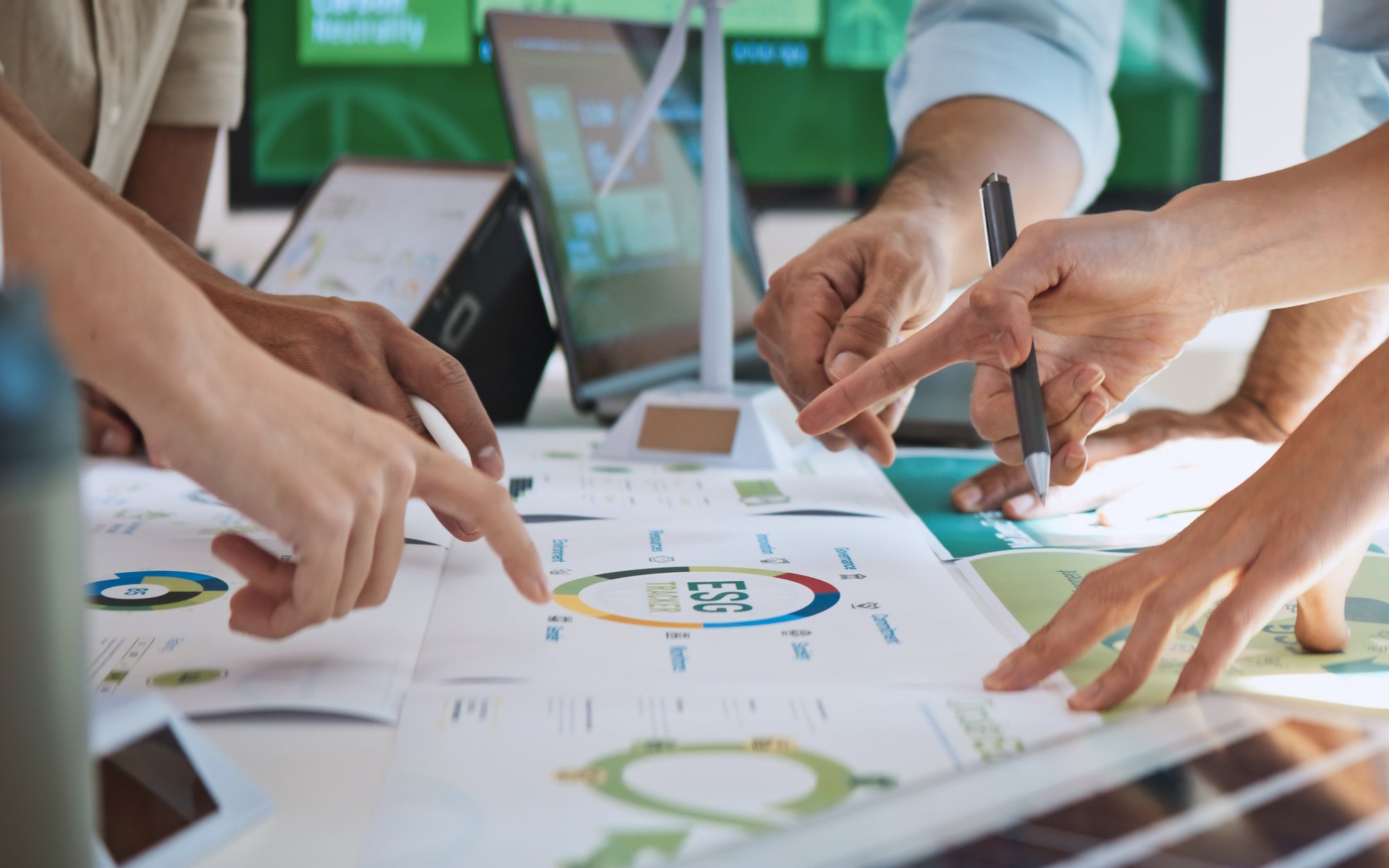 Multiple hands pointing at a technical diagram or blueprint spread out on a table. Electronic devices and screens are blurred in the background.